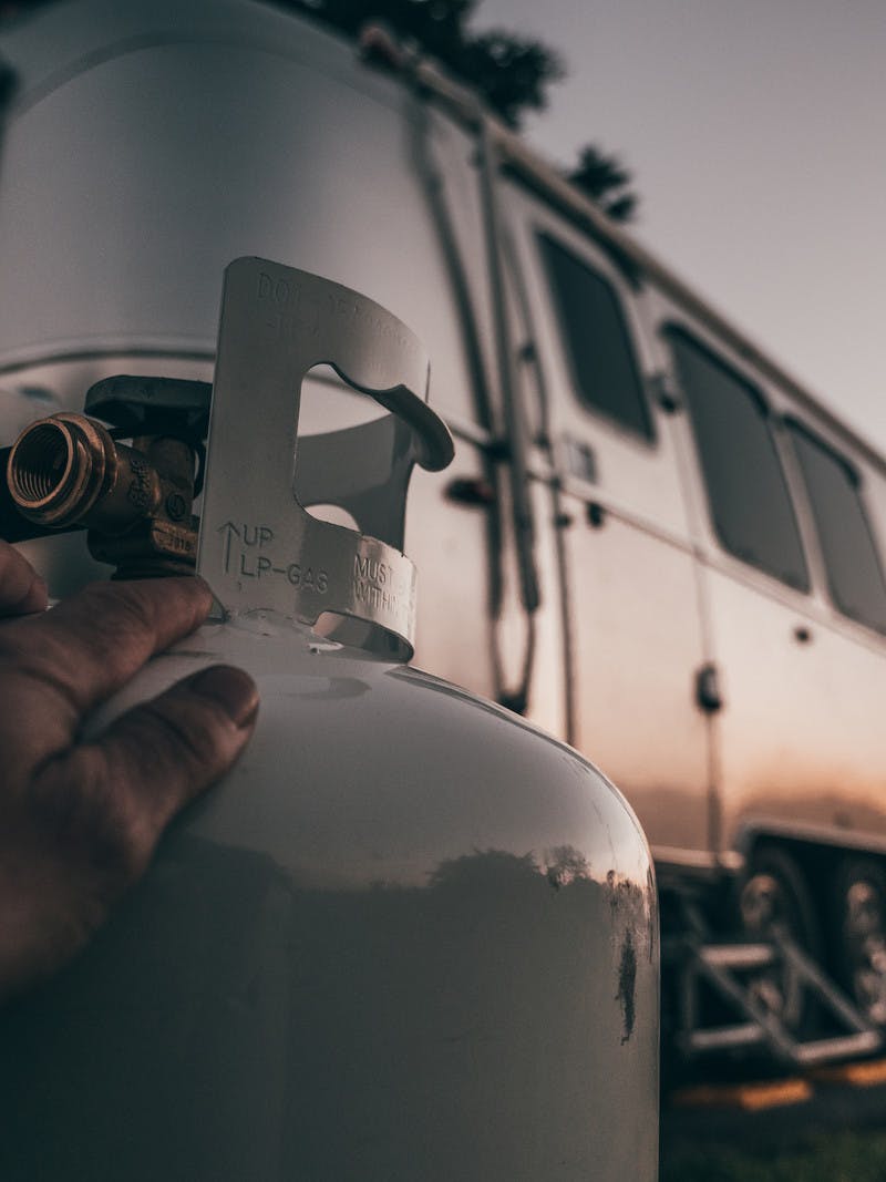 A close up shot of a hand on a propane tank with the sun setting on an RV in the background. 