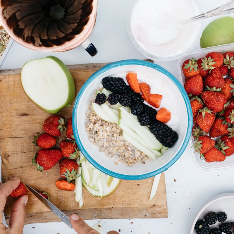 A top down picture of a white counter. On it, a hand cuts strawberries on a wood cutting board. A pint of strawberries sits nearby. A halved apple sits nearby.