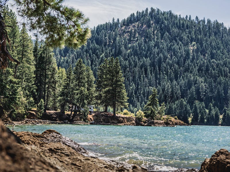 Coast line of Rimrock Lake, sparkling water, RV on lake edge, lots of evergreen tress around.