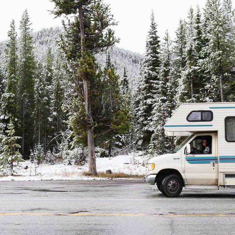Class C motorhome driving down wet road with snowy pine trees.