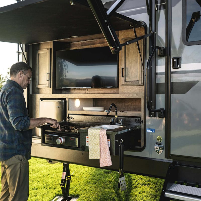 A man grills food on the outdoor grill and entertainment system outside of a Venutre RV.