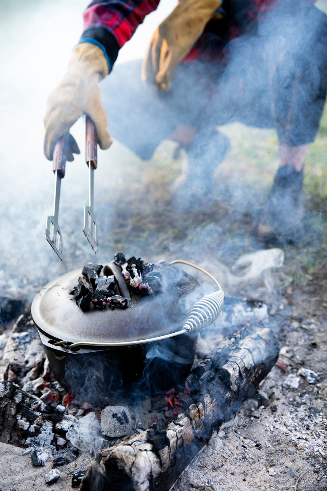 A dutch oven cooks over an open fire with coals on top of the lid. 