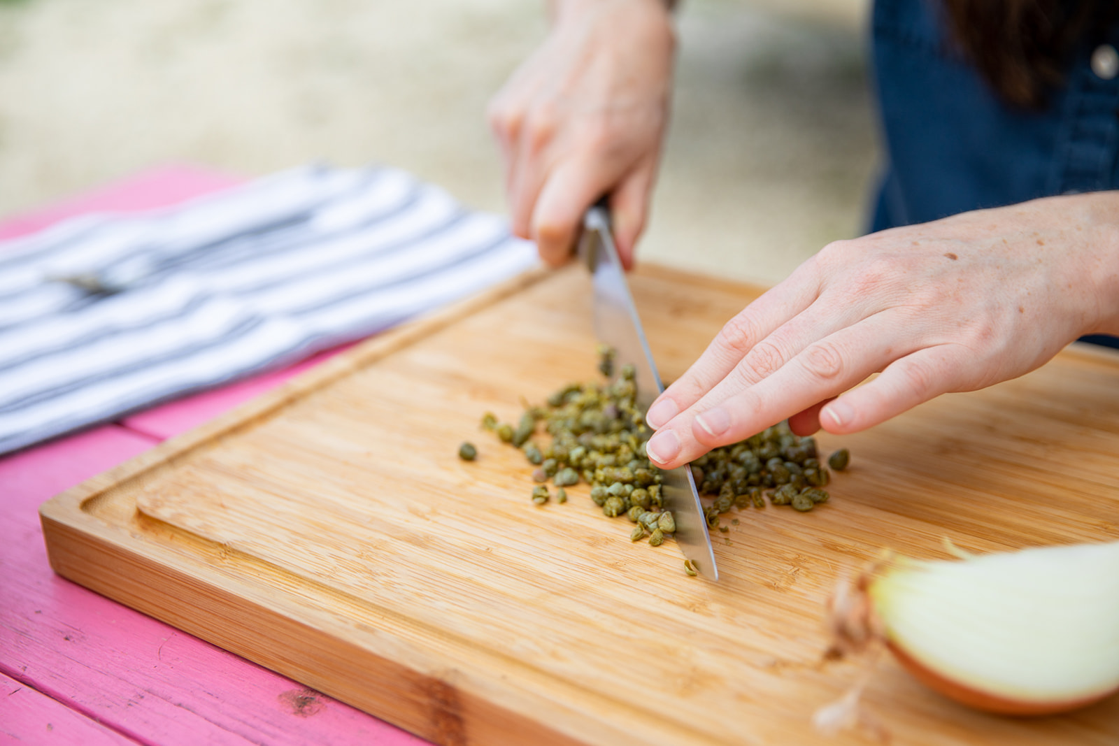 Hands holding a knife that chops capers on a wooden cutting board. 