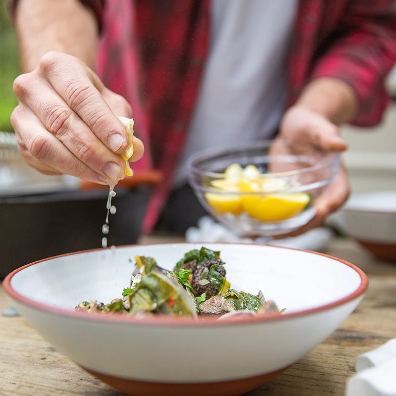 Hands squeezing lemons over white bowl of cooked clams with parsley.