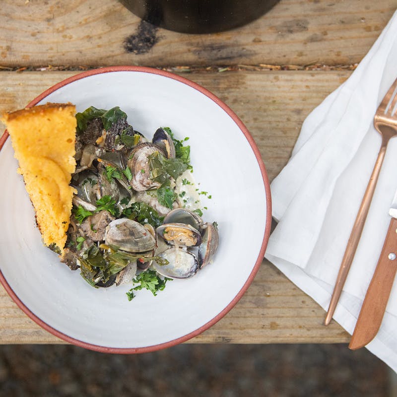 Bowl of cooked clams with parsley on a white plate, a golden slice of cornbread, and gold utensils on a white napkin.