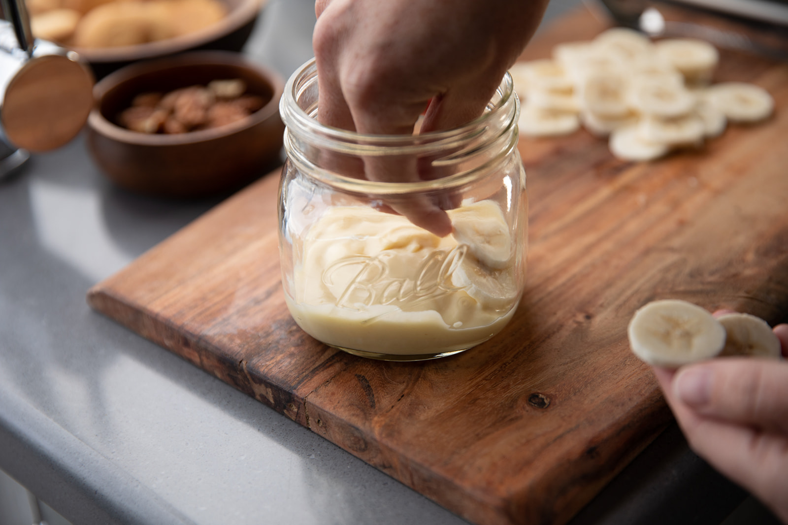 A hand layering banana slices on top of a layer of pudding in a glass jar. 