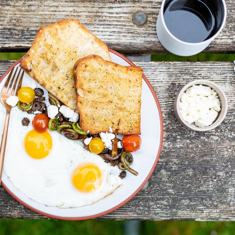 A finished breakfast plate with 2 sunny-side eggs, vegetable mix topped with feta cheese, and 2 pieces of toast, with a mug filled with coffee on a picnic table.