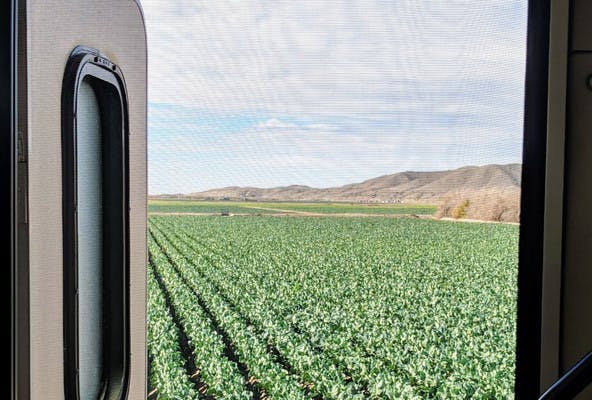 View outside the front screen door of an RV, looking out at a farm field, with brown mountains in the background.