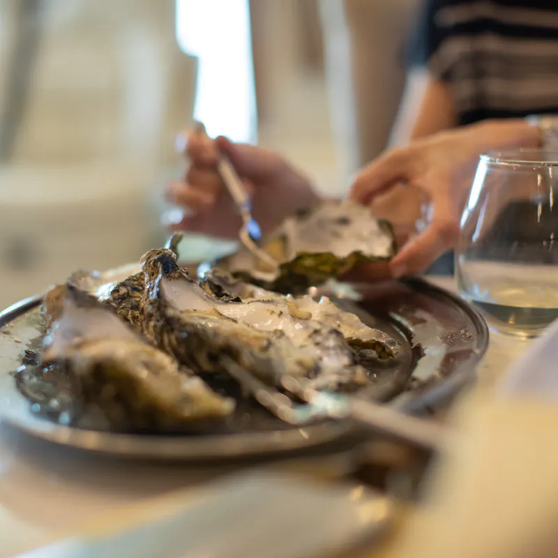A close-up of fresh oysters on a metal platter with a person preparing to eat one in the background.