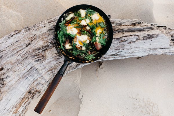 A cast iron skilled filled with cooked greens, bacon and eggs, balanced on a piece of driftwood on the beach.