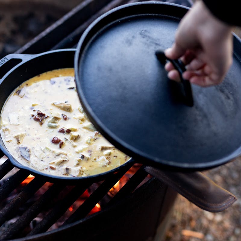 Bacon artichoke frittata being cooked over a fire, while woman prepares to put lid on pan.
