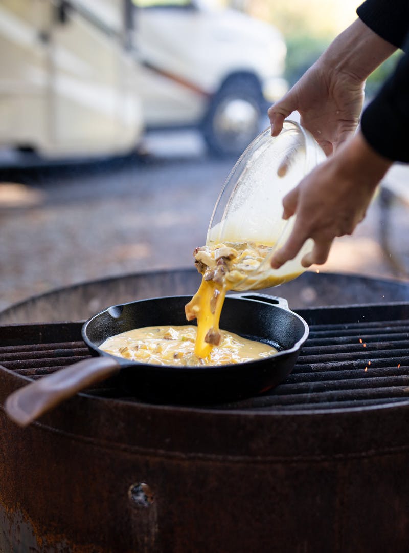 Woman pouring egg mixture from a bowl onto the pan over a fire.