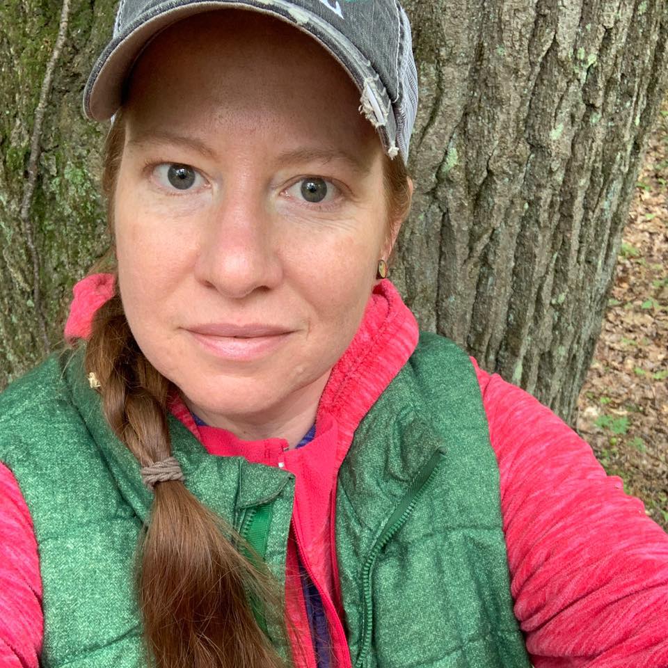 A selfie of Amy Lundquist-Stachowiak sitting in front of a tree wearing a hat and green vest