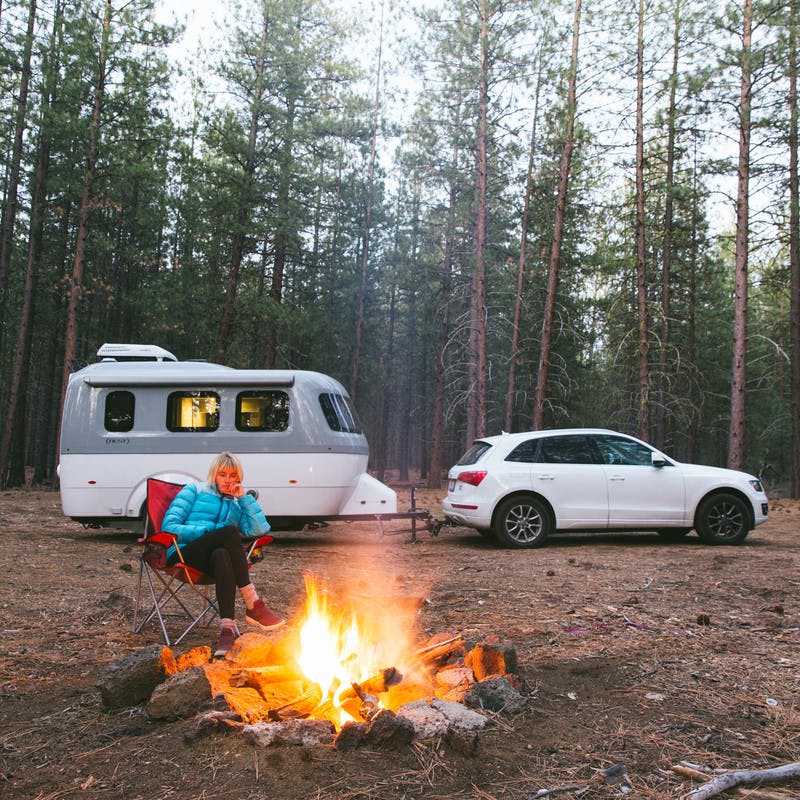 A blonde woman dressed in a winter hat, pants, and gym shoes, sits by a campfire with her white car and Airstream nest parked behind her.