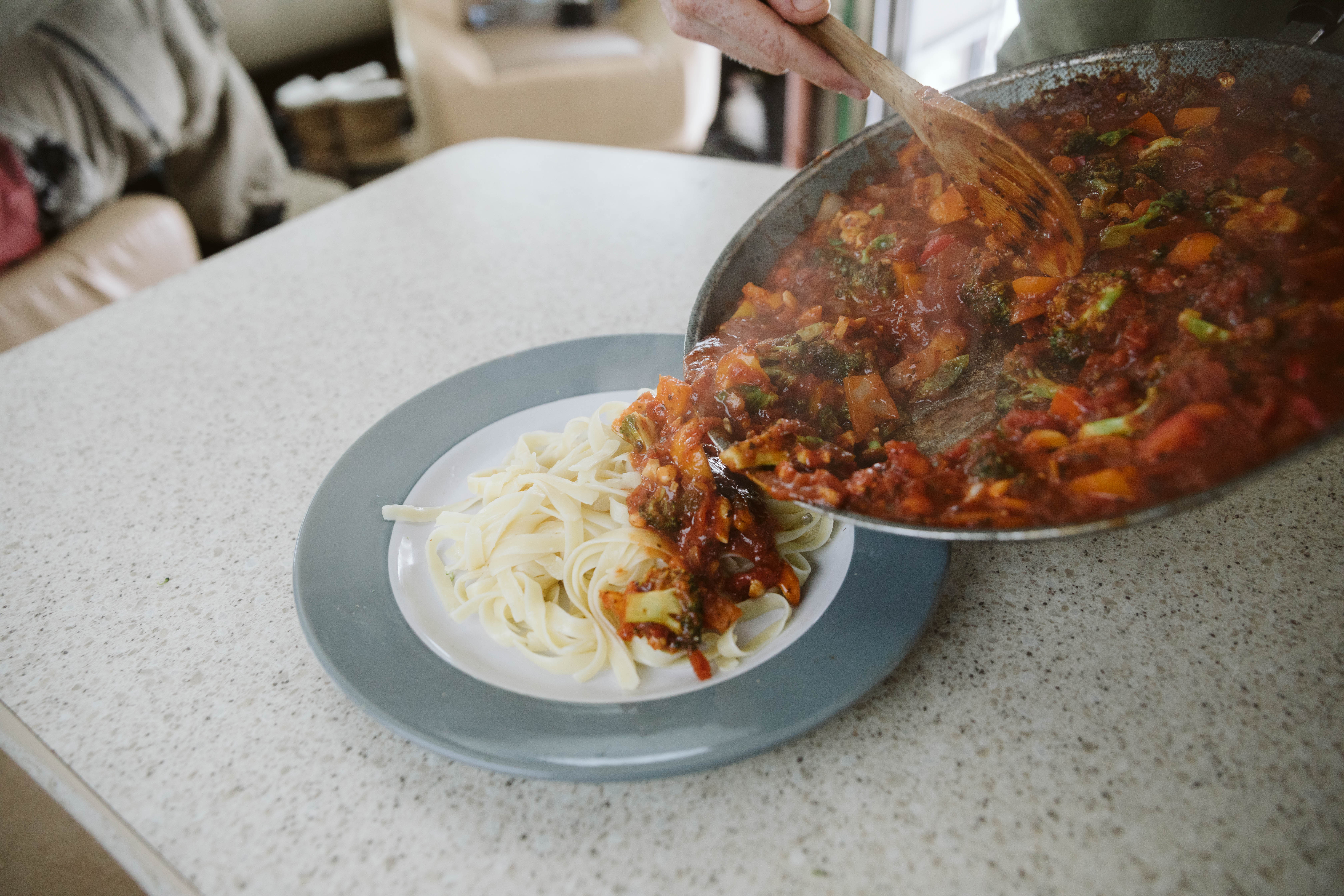 Pan pouring vegetable and red sauce mixture onto plate of pasta