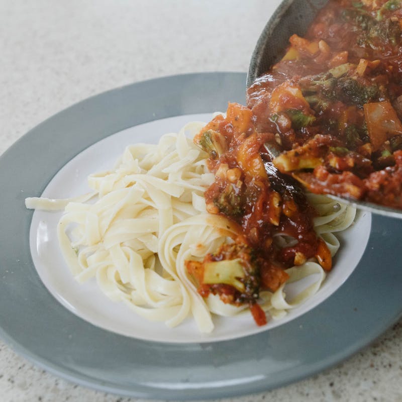 Pan pouring vegetable and red sauce mixture onto plate of pasta