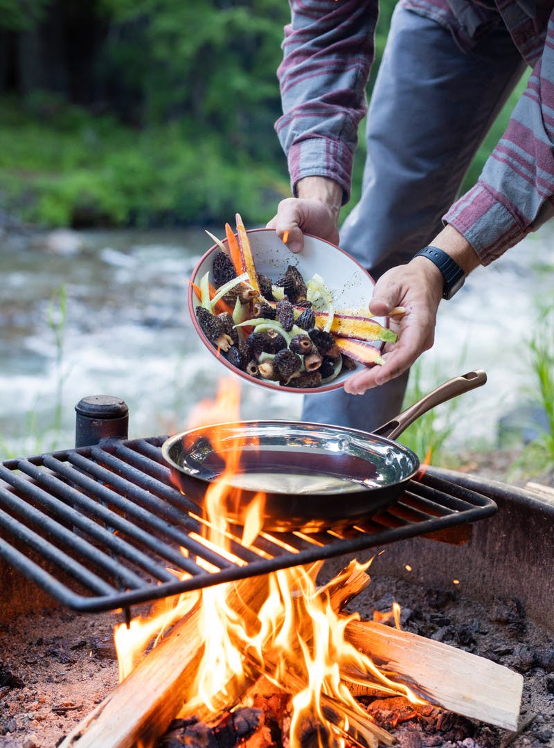 Man in flannel drops bowl of carrots, morels and fennel into frying pan over campfire.