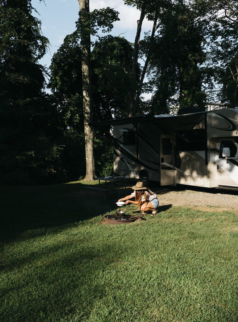 A shot of Sarah cooking over a small flame in the outdoors, surrounded by trees and grass and sky. 