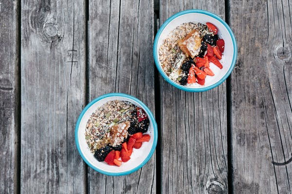 White bowls with a sky-blue rim sitting on a gray, aged wood picnic table. The bowls hold servings of overnight oats topped with colorful stripes of berries, chia seeds and honeycomb.