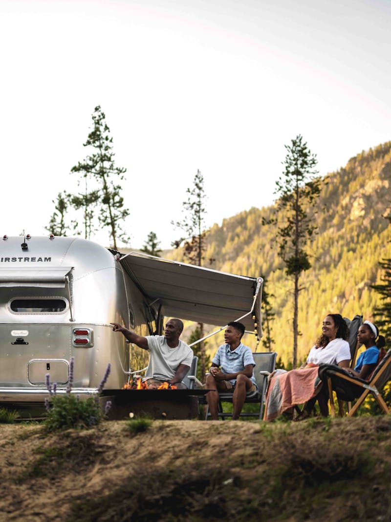 A family gathers around a fire at a campsite with a Airstream Flying Cloud.