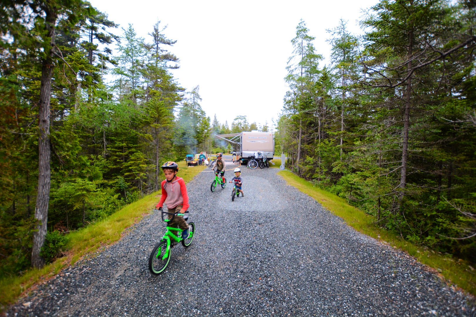 Three young boys riding bikes at a tree-lined campsite.