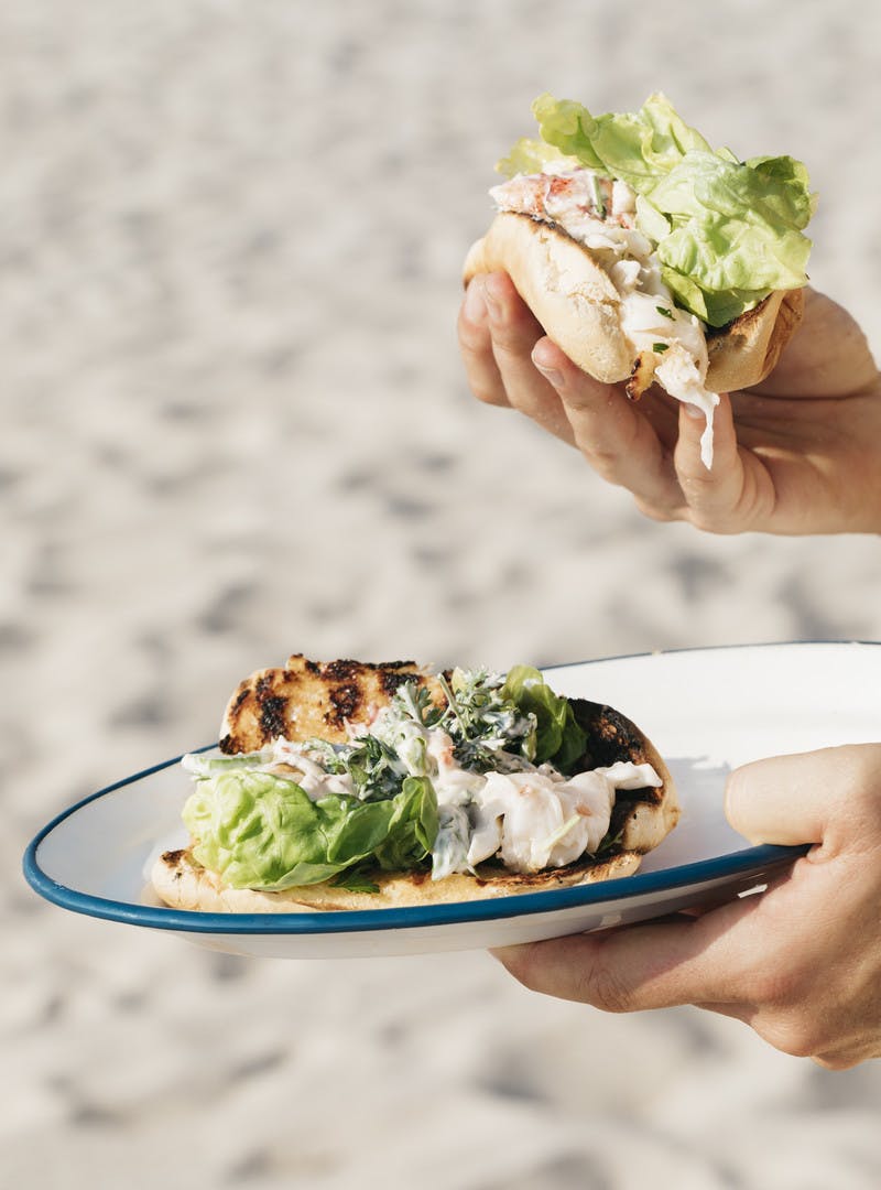 A hand holding an assembled lobster roll as well as another hand holding a plate with another lobster roll on it. 