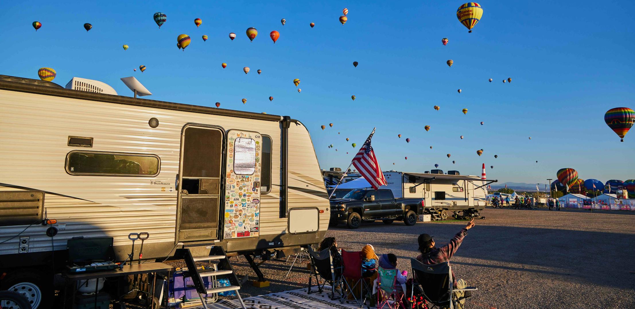 The Brian and Surna Barton family sitting at their campsite with their Coleman Travel Trailer while looking at hot air balloons at the Albuquerque Balloon Fiesta