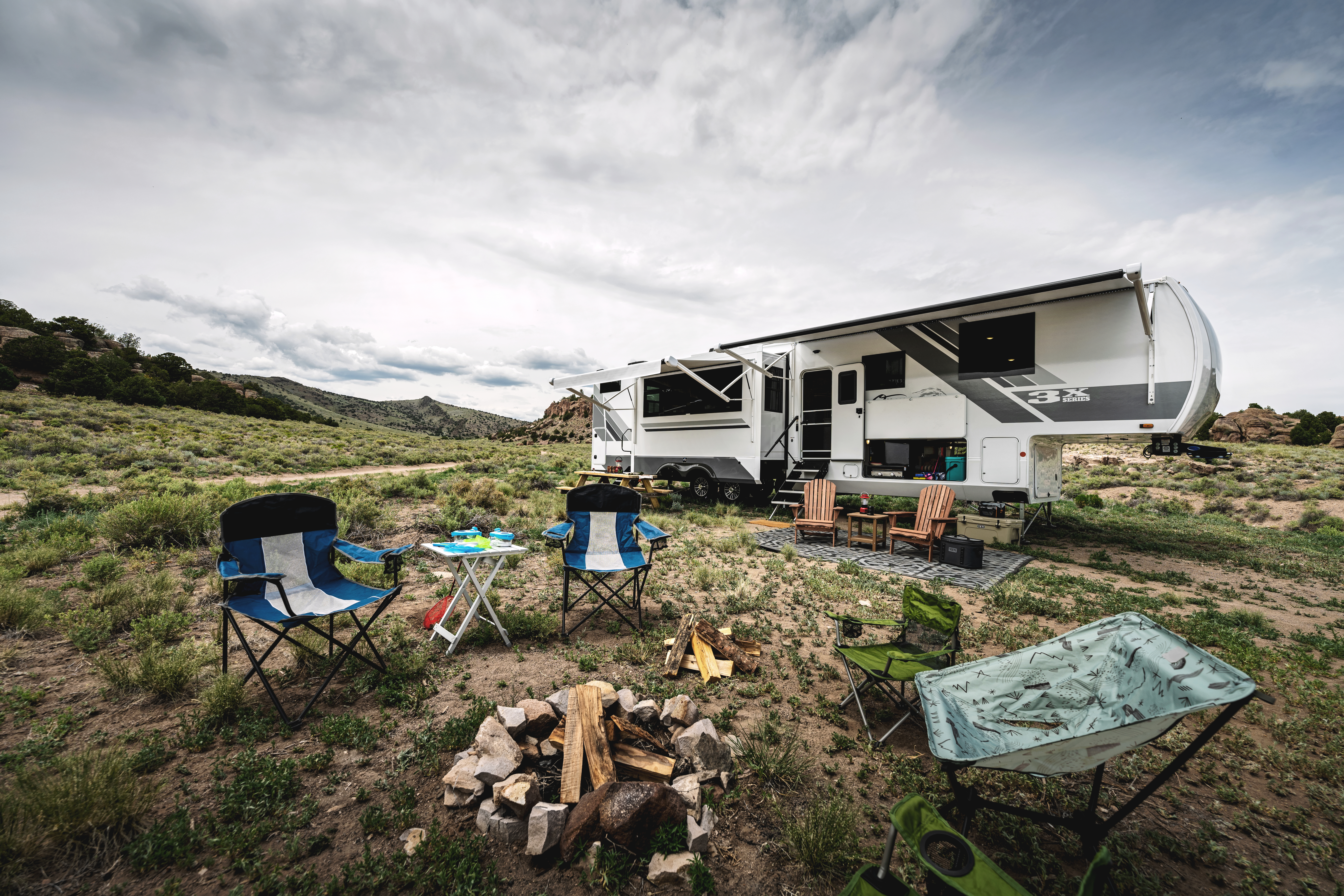 A firepit surrounded by campchairs in front of an Open Range fifth wheel in the mountains. 