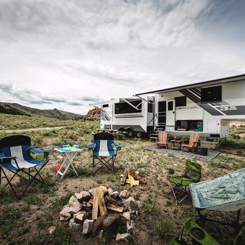 A firepit surrounded by campchairs in front of an Open Range fifth wheel in the mountains.
