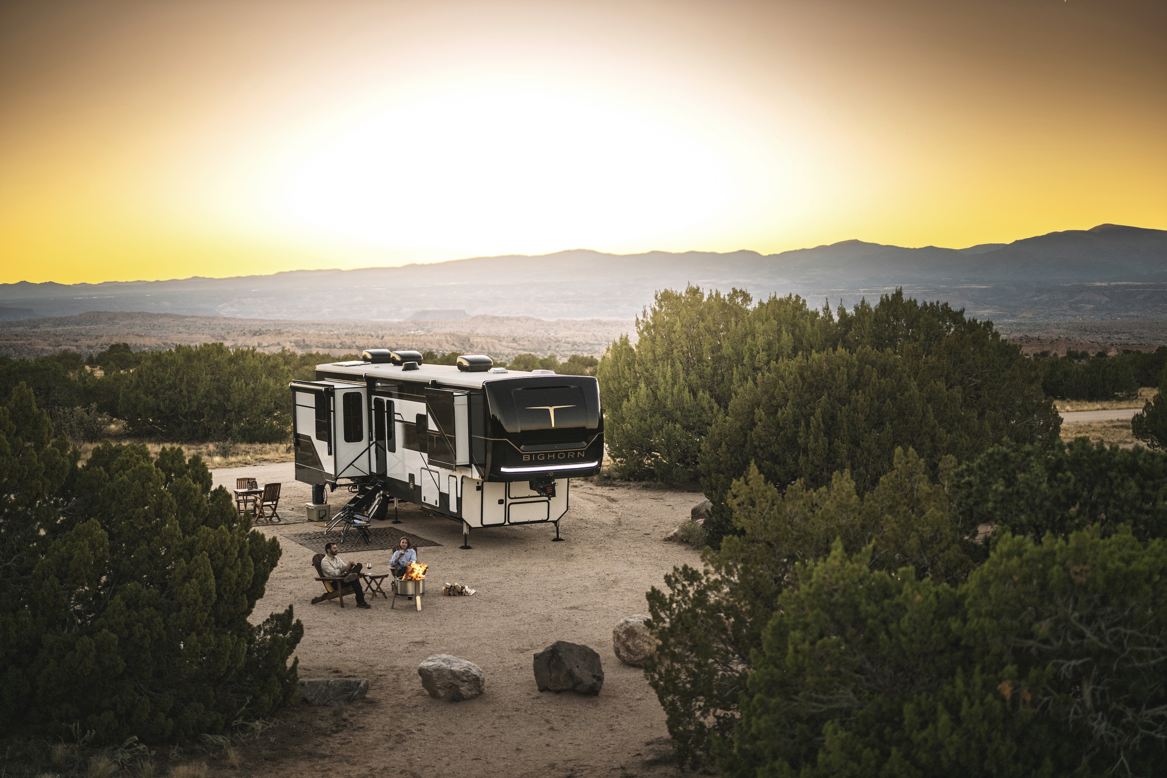 A couple sits around a campfire drinking wine as the sun sets behind them and their Heartland Torque fifth wheel.