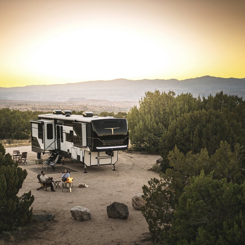 A couple sits around a campfire drinking wine as the sun sets behind them and their Heartland Torque fifth wheel.