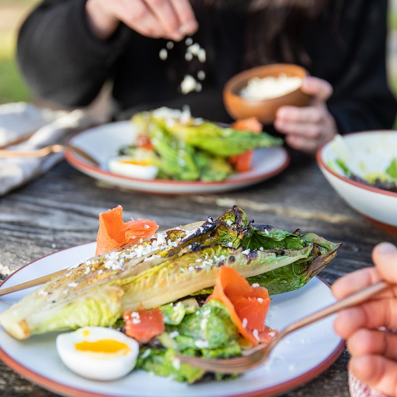 Woman sprinkles fresh shaved parmesan cheese on her caesar salad while man holds fork, preparing to eat his dish.