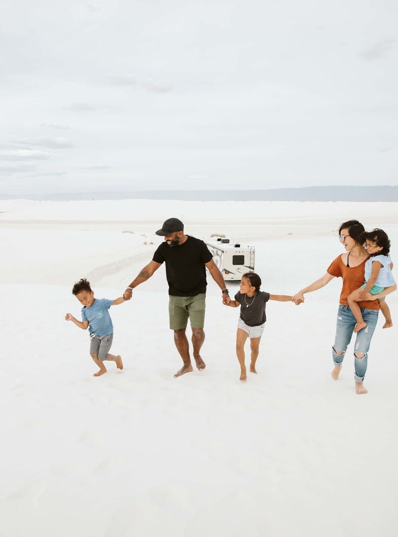 Family of five, holding hands, carry child, as they walk through white sand.