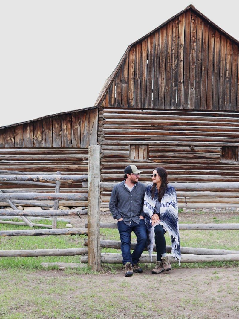 Juli & Jordan Cote stand in front of wooden fence and log barn, Juli with sunglasses and blanket wrapped around her and Jordan with a baseball hat on.