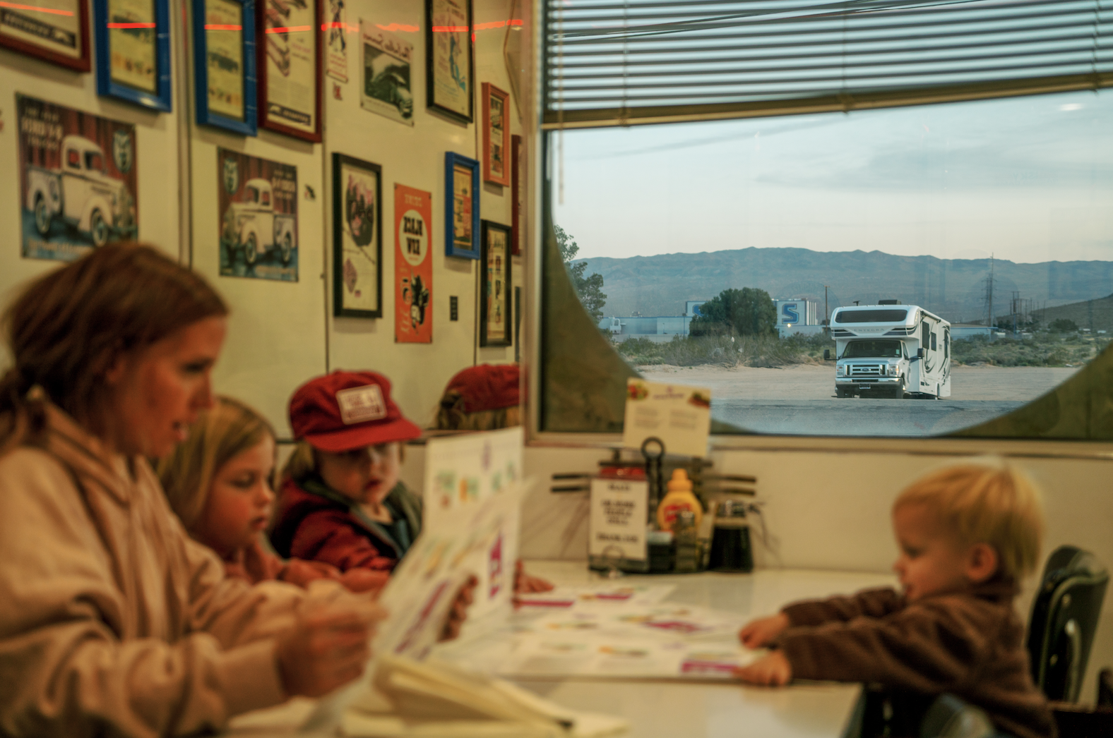 Mom sits at a diner table with three young kids reading menus while class c motorhome sits outside window 
