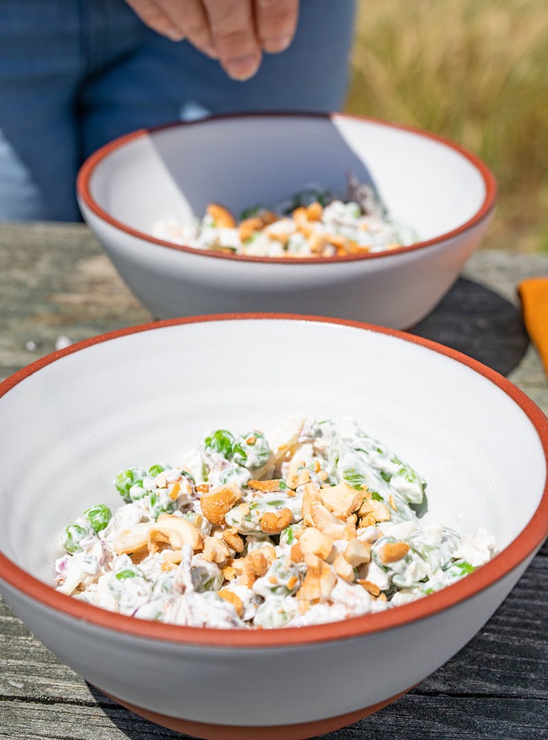 Two white bowls, with hand sprinkling chopped cashews on pea salad.