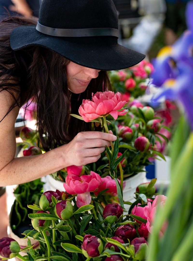 Aubrey in a floppy black hat stops to smell pink and purple flowers.