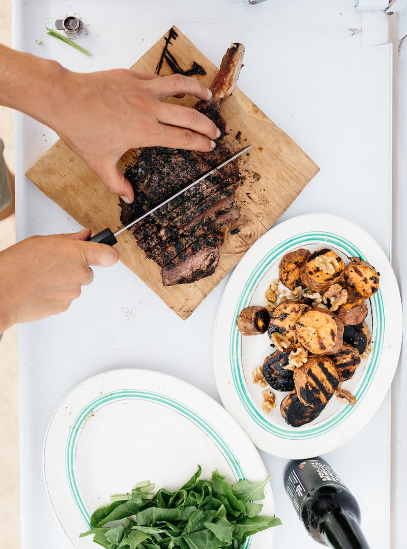 Two white dishes, one with a pile of greens and another with the sweet potato rounds, cooked. A hand slices the steak on a cutting board.