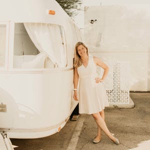 Kristiana Spaulding, in a cream-colored dress and flats, leaning against her Airstream RV in a parking lot, smiling at the camera.