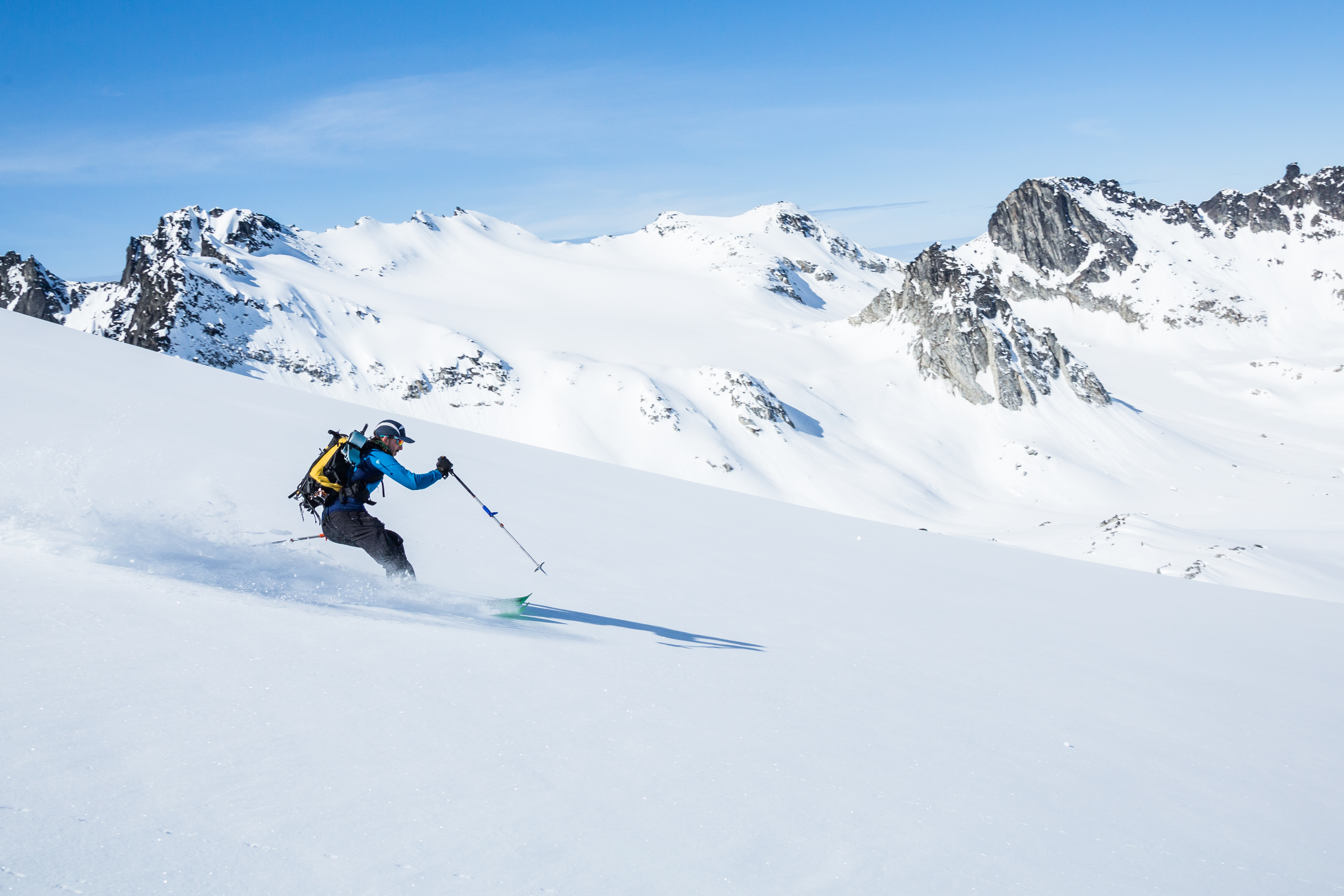 Man with yellow backpack skis down snowy mountain with jagged mountain peaks in the background