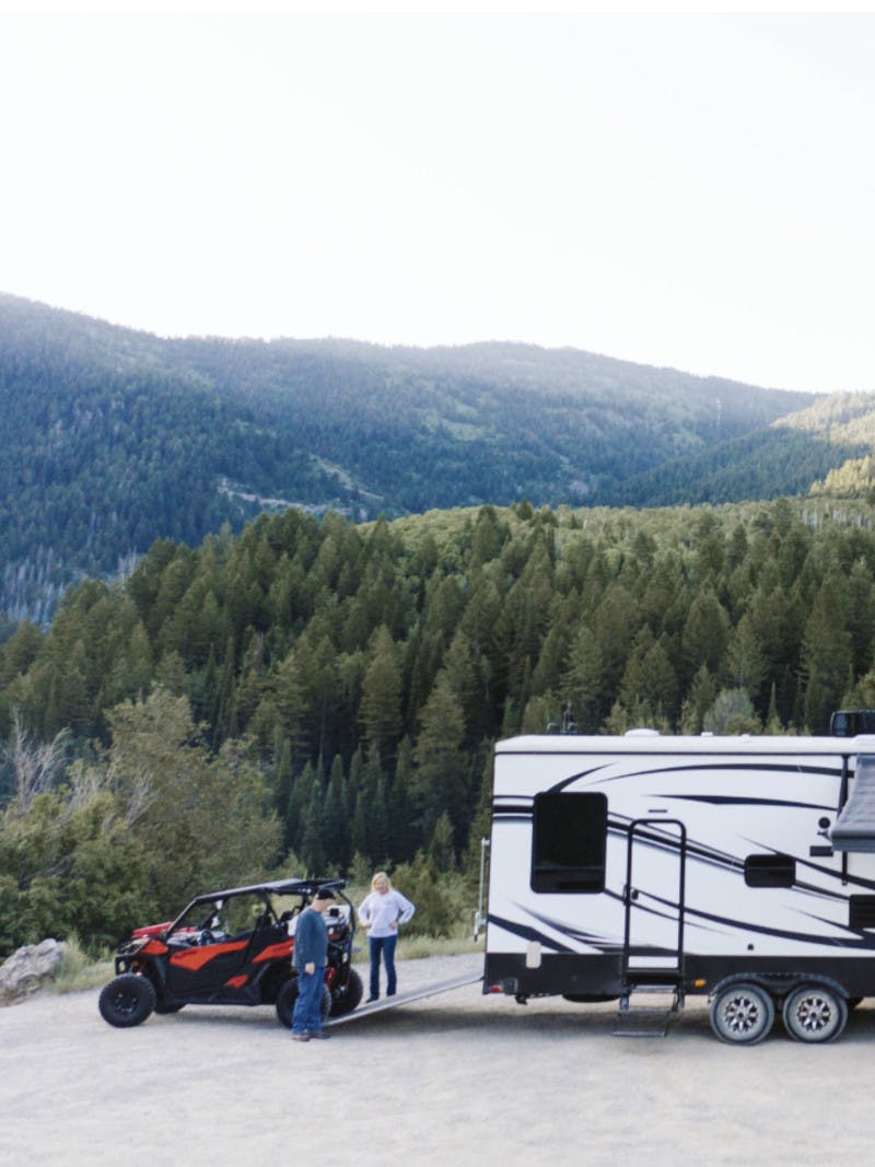 Steve & Suzanne Talbot standing next to an ATV being unloaded from a Toy Hauler RV hitched to a truck with a mountain backdrop. 