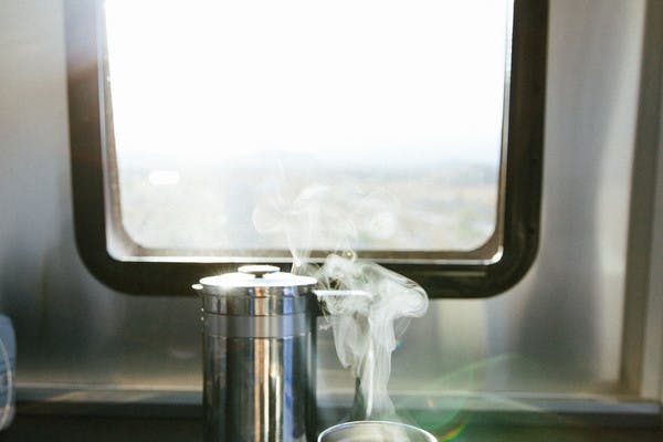 Steaming cup of coffee, next to a metal French press, in front of an RV window.