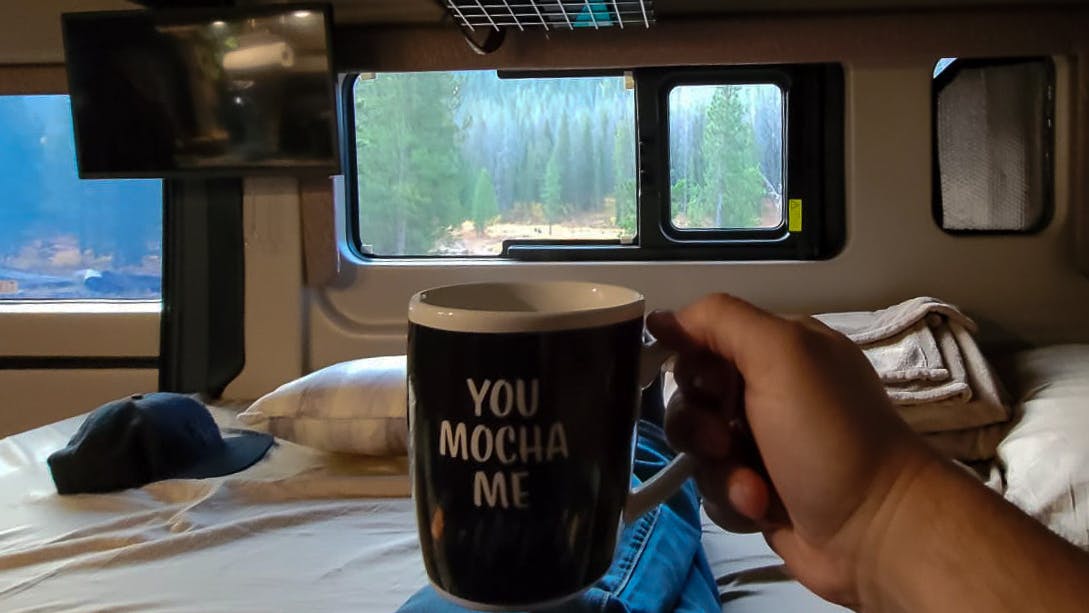 Rocio holding up a cup of coffee with a view of a National Forrest out of her camper van window