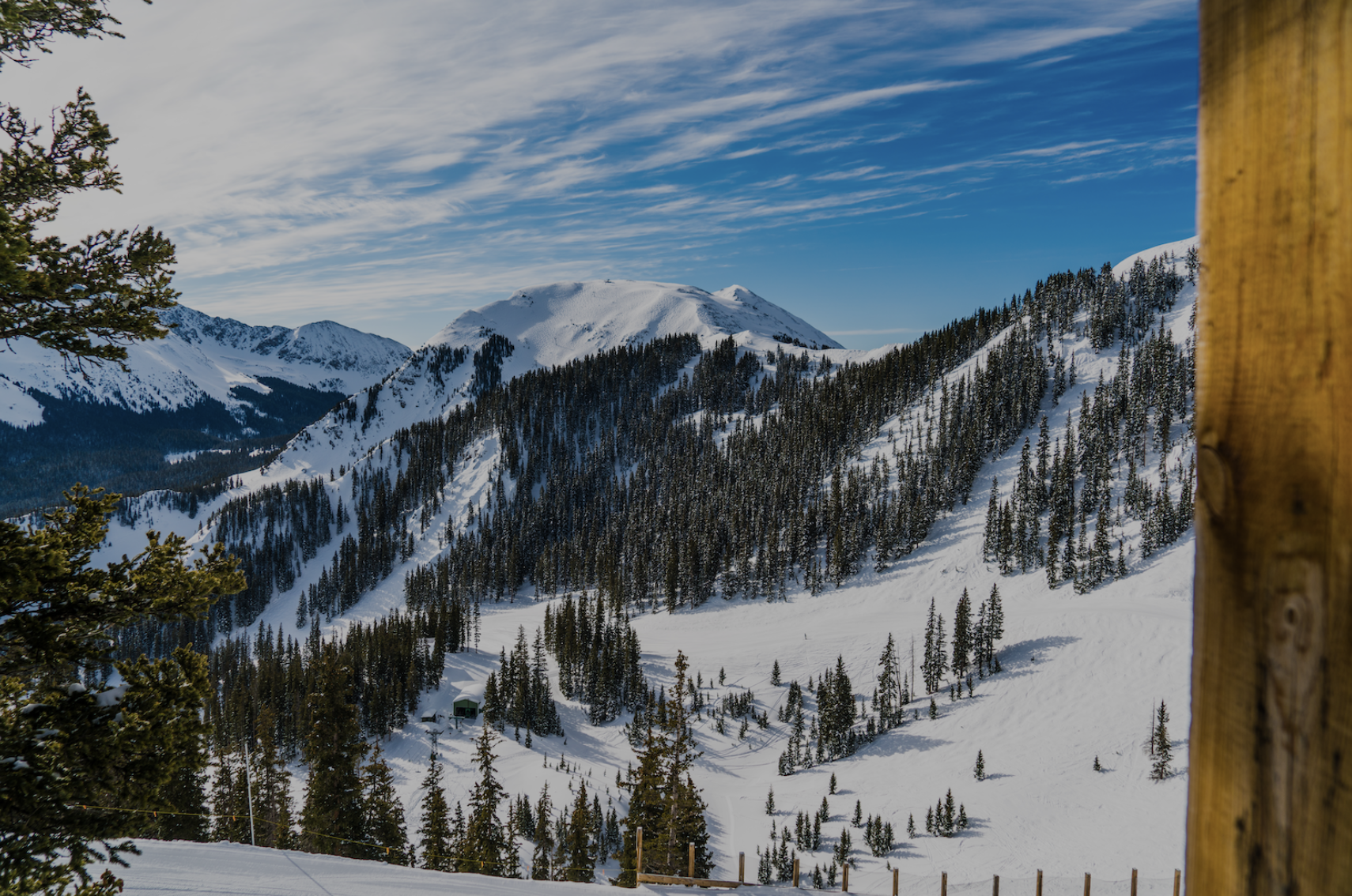 Looking out next to a wooden pole onto blue skies and snowy mountain range covered in trees