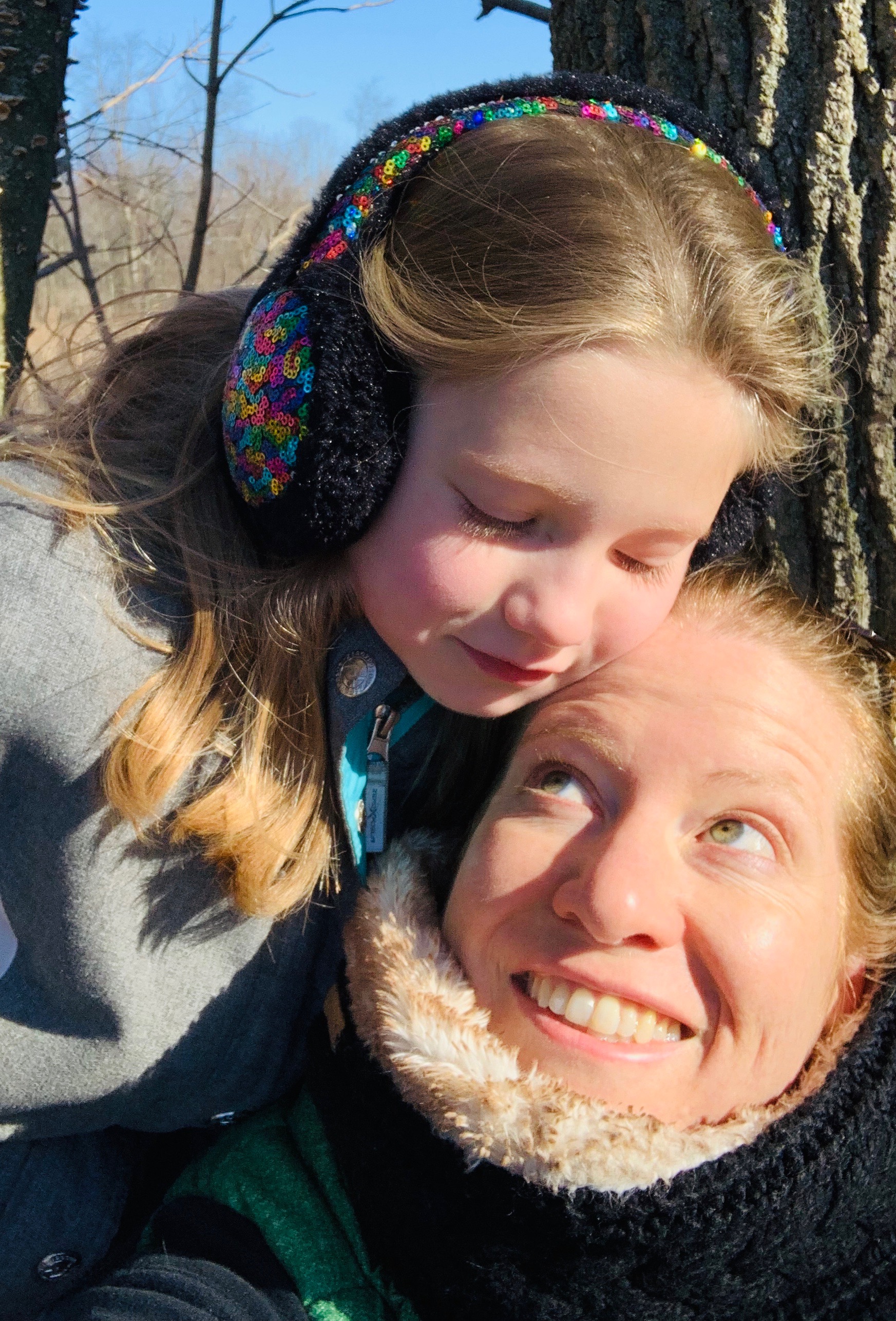 Young girl rests cheek on mother's forehead as mother gazes up and smiles