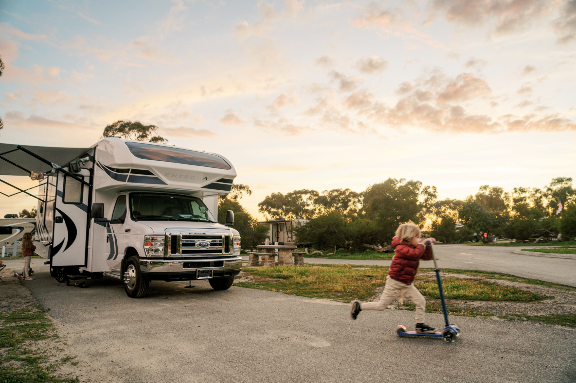 Little boy rides scooter down pavement with class c motorhome in background at sunset