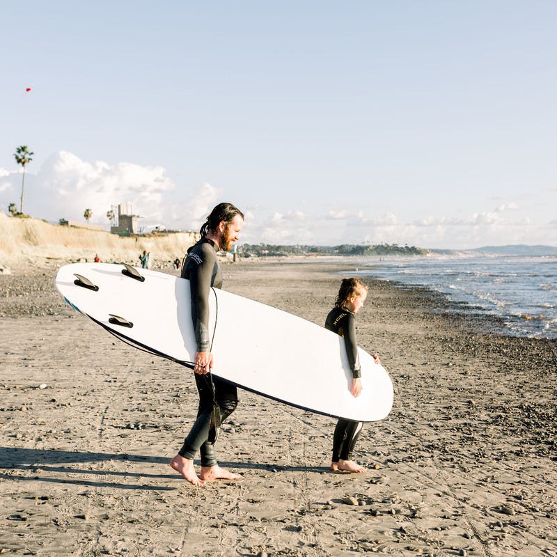 Father and daughter carrying a surfboard on the beach towards the water.