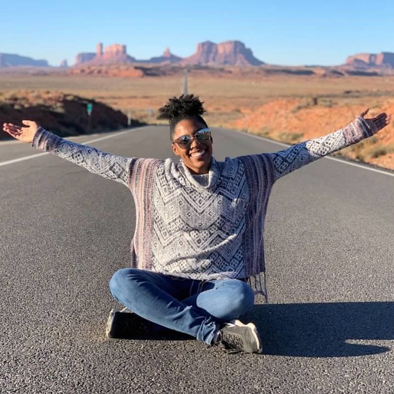 An African American woman sits in the middle of the road with her arms spread wide, with Monument Valley in the far background.