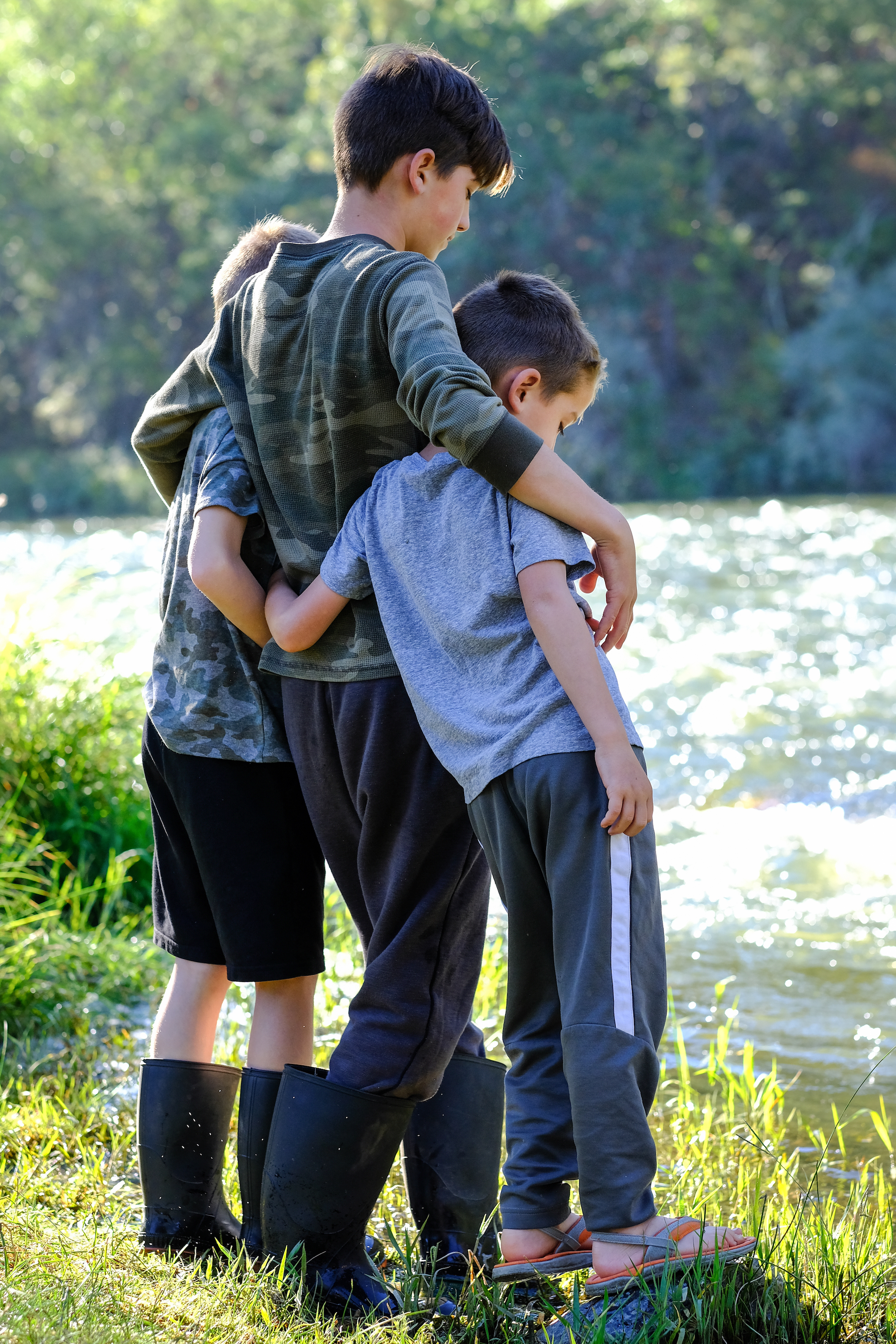 Three young boys stand arm in arm by a river.