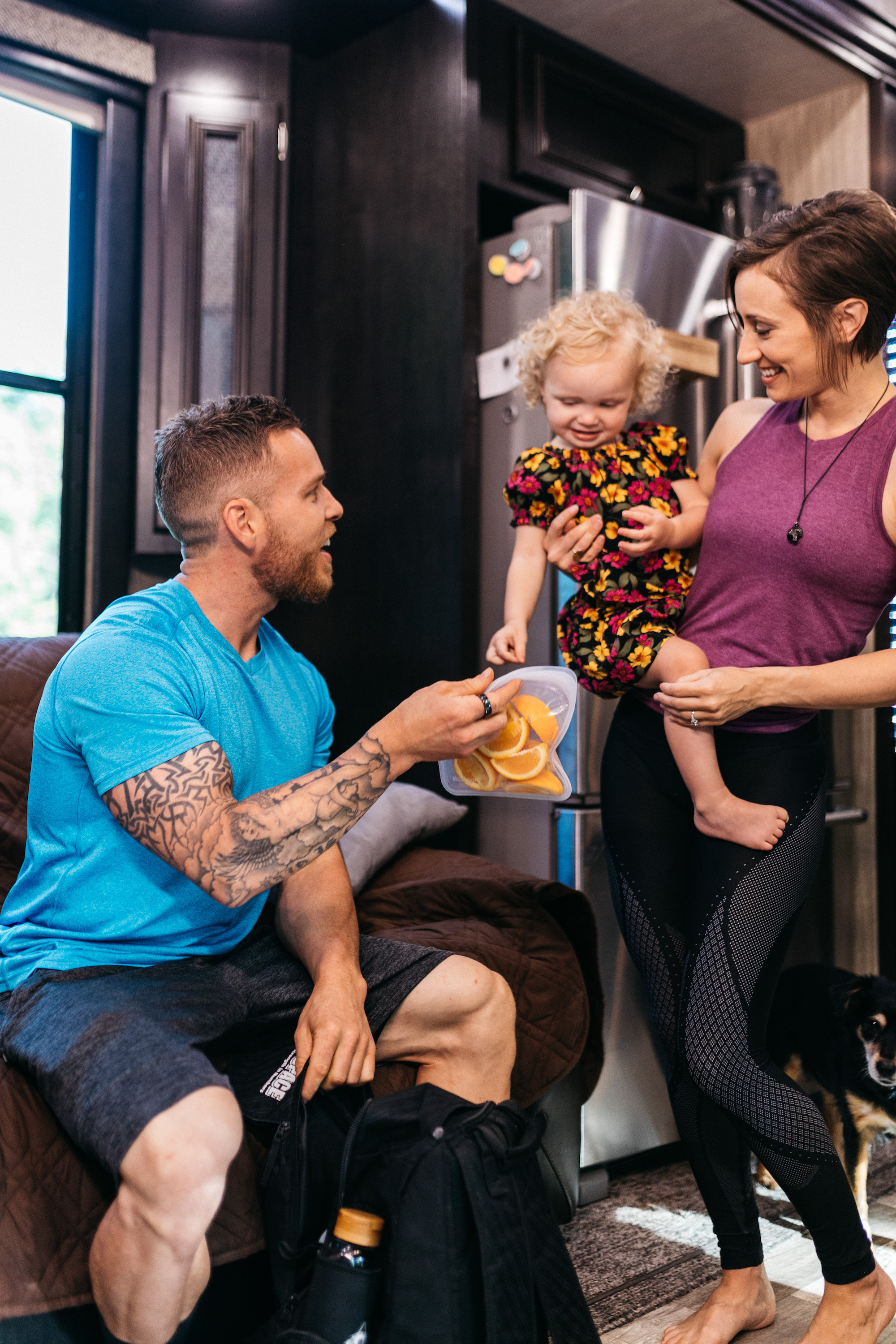 A man holds a baggie of cut orange slices up so his daughter can take one.